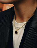 Gold necklace with a square pendant worn by a person in a dark jacket and white shirt.