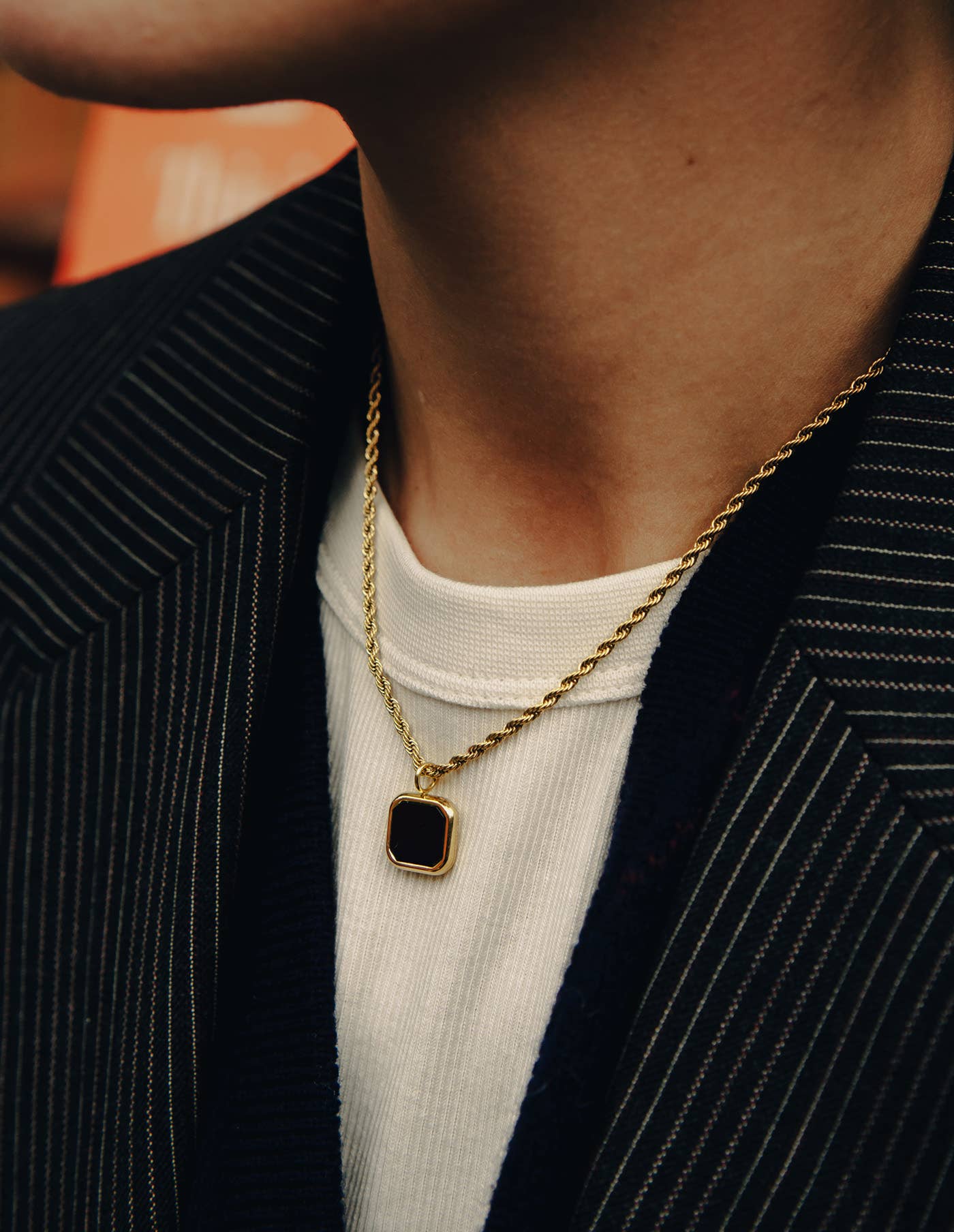 Gold necklace with a square pendant worn by a person in a dark jacket and white shirt.